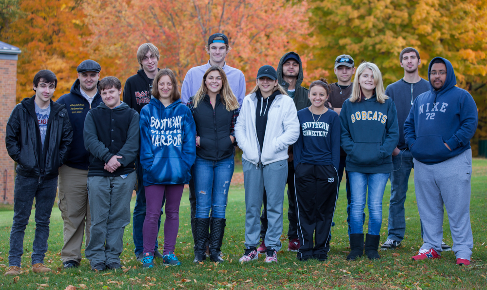 group of students standing outside in the grass, fall foliage behind them