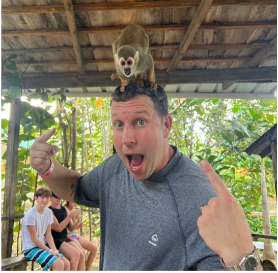 A Flo balances a small monkey on his head while pointing at it with both hands and making a face of amazement. Wow!